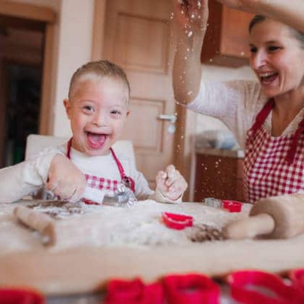 A laughing handicapped down syndrome child and his mother with checked aprons indoors baking in a kitchen, having fun.